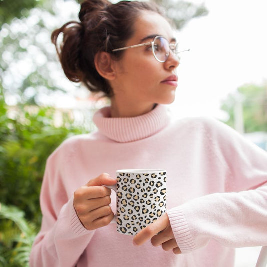 Woman wearing sunglasses and a light pink turtleneck holds a white mug with a black leopard-print pattern outdoors.