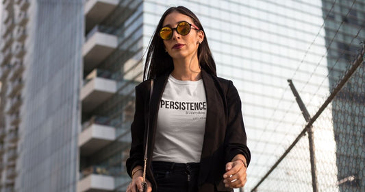 Woman in black blazer and sunglasses wearing white Persistence Is Everything graphic tee by Leela's Ink on escalator