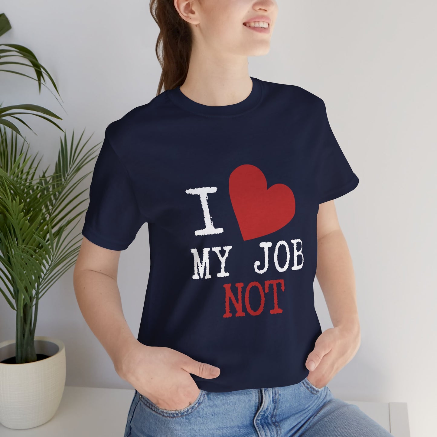 A woman wearing a navy blue graphic t-shirt with the text 'I [red heart] MY JOB NOT' in white and red, paired with blue jeans, in a brightly lit indoor setting with a potted plant on the left.