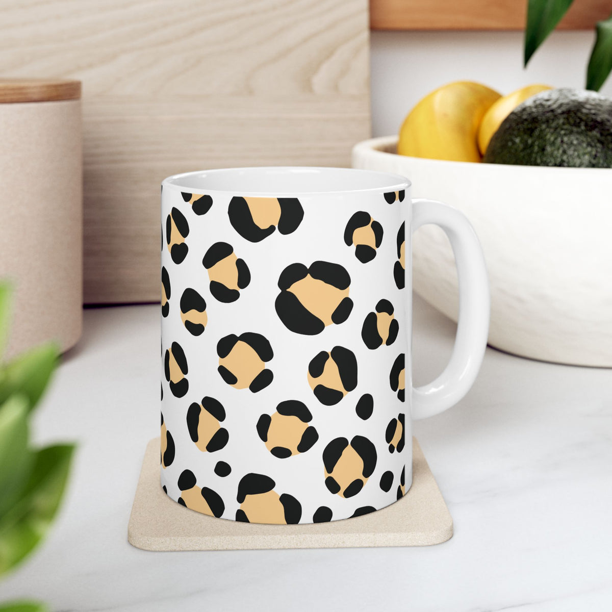 A white ceramic mug with a black and tan leopard print pattern, resting on a beige coaster on a white marble countertop with a blurred kitchen background.