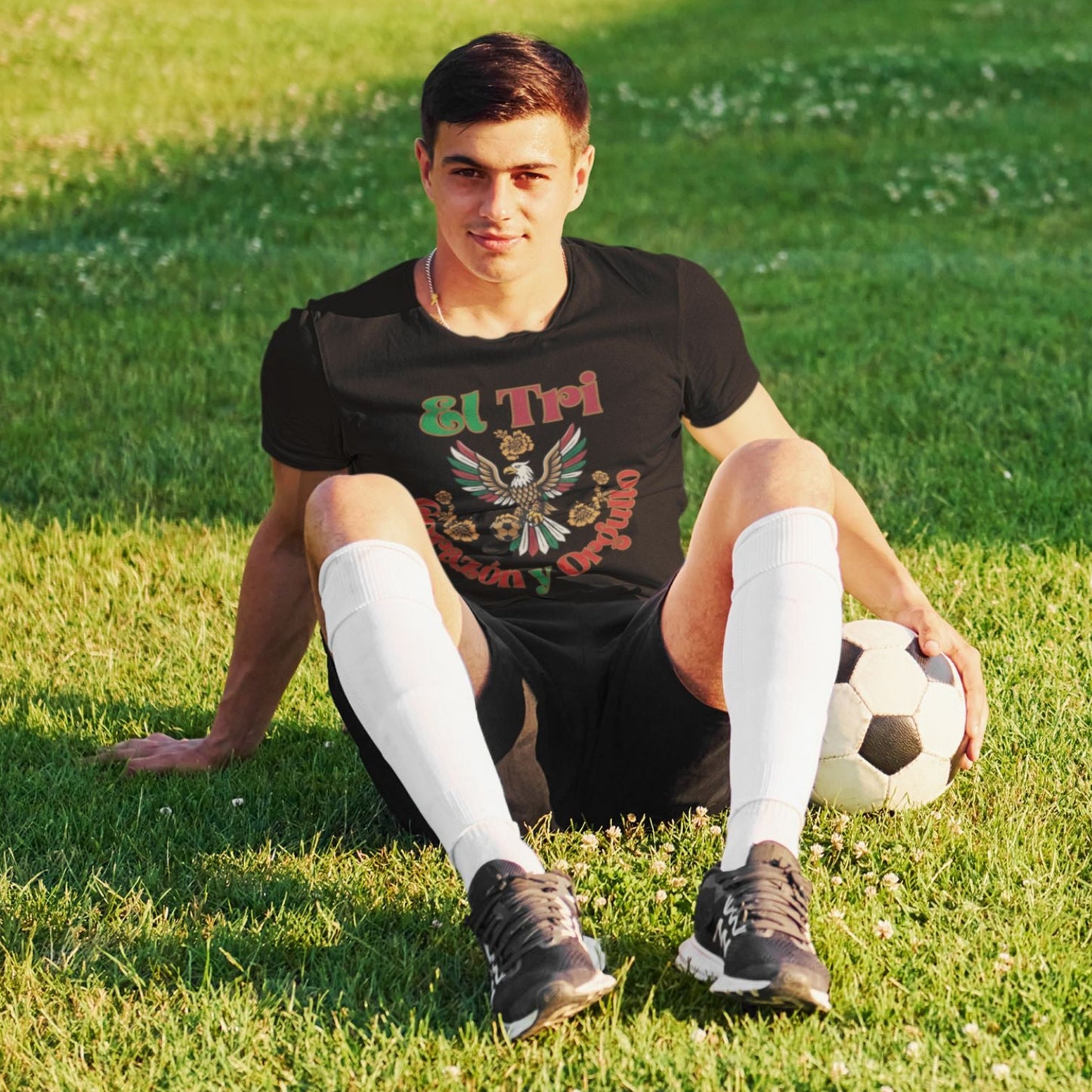 Man sitting on grass in black El Tri Corazón y Orgullo tee with soccer ball and cleats, outdoor lifestyle shot
