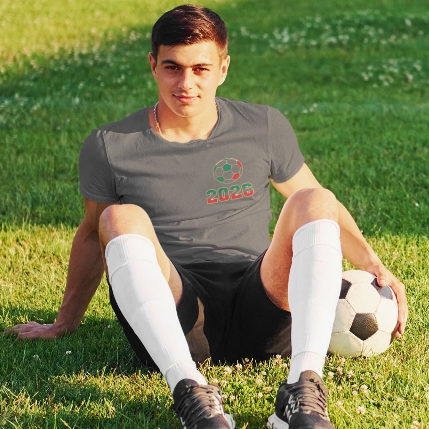 Man sitting on grass in charcoal El Tri soccer tee with chest logo, soccer ball and cleats, outdoor lifestyle shot
