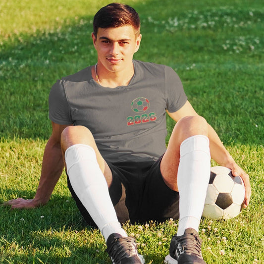 Man sitting on grass in charcoal El Tri soccer tee with chest logo, soccer ball and cleats, outdoor lifestyle shot
