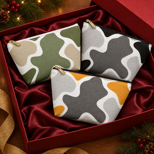 Three abstract camo accessory pouches in sage green and tan, black and gray, and mustard and gray, arranged in a red velvet gift box with gold-tone zipper pulls.