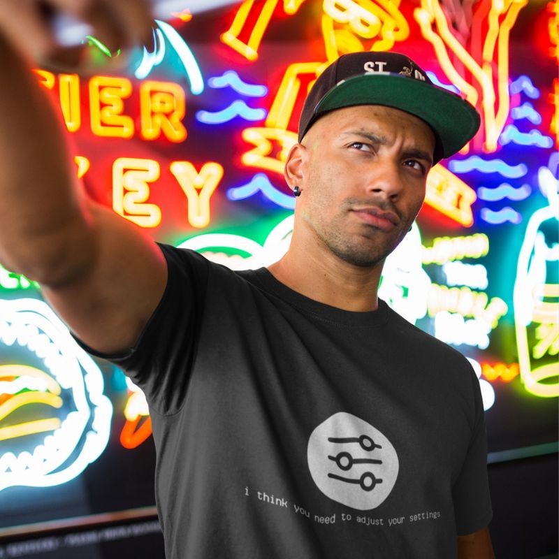 Man taking a selfie in front of neon signs wearing a black I Think You Need to Adjust Your Settings tech graphic t-shirt.