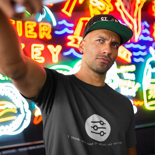 Man taking a selfie in front of neon signs wearing a black I Think You Need to Adjust Your Settings tech graphic t-shirt.
