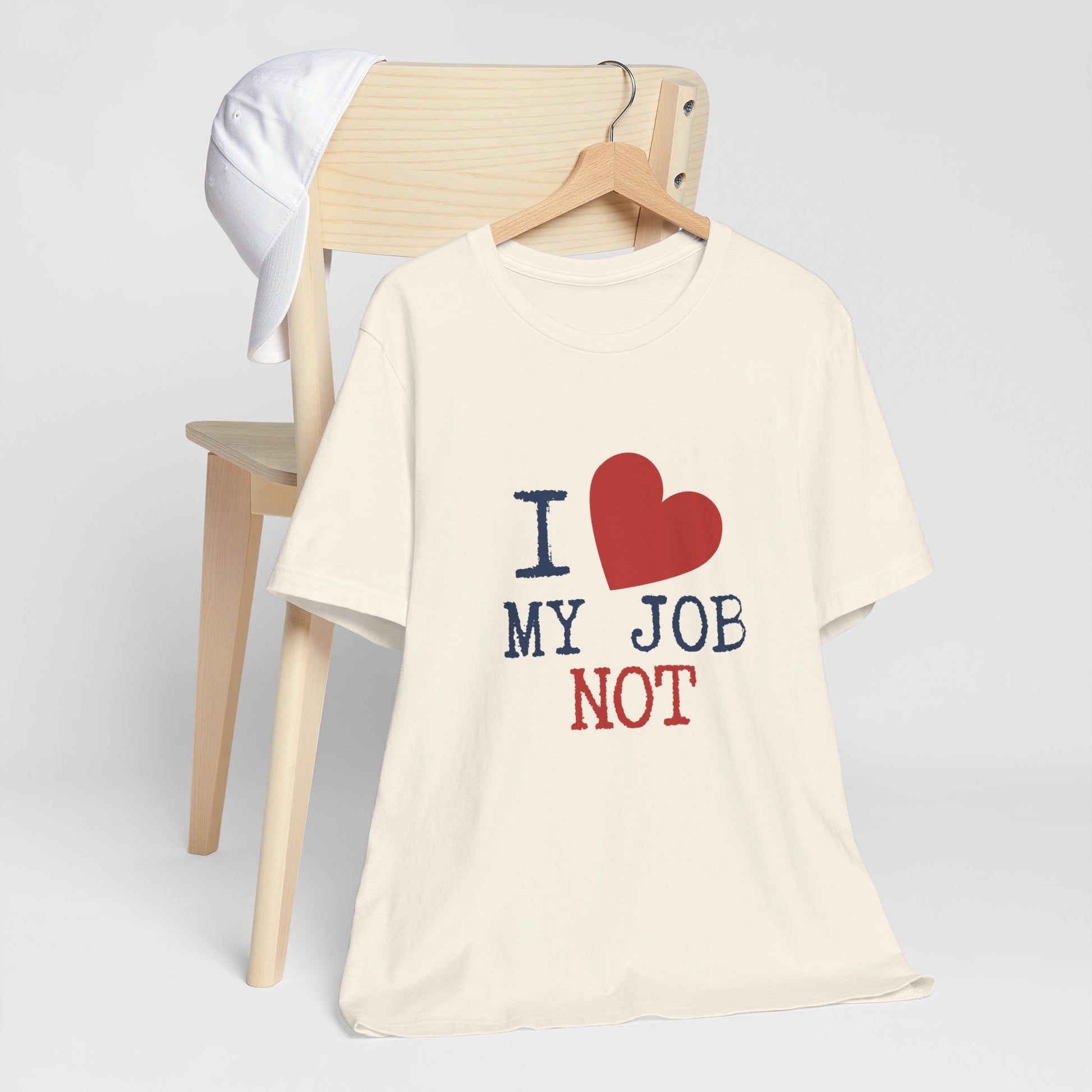 An off-white short-sleeve t-shirt on a wooden hanger, styled in front of a light wood chair with a white cap hanging off the backrest. The shirt features a graphic print that reads 'I' followed by a red heart, 'MY JOB', and 'NOT' in navy blue and red text.