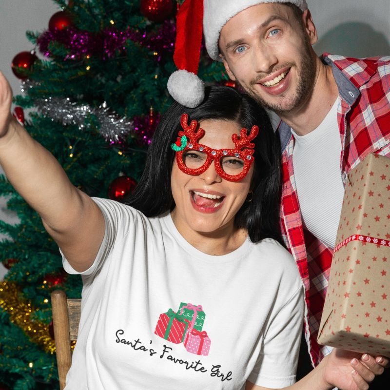 A smiling woman and man pose together in front of a decorated Christmas tree. The woman wears a white graphic T-shirt with a red and green gift illustration and the text 'Santa's Favorite Girl,' along with red reindeer glasses.