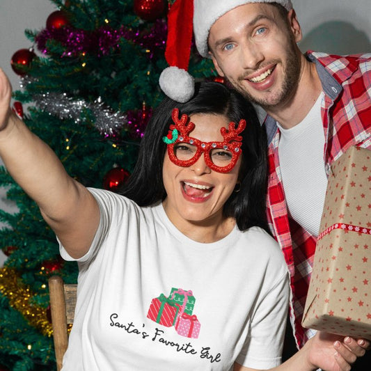 A smiling woman and man pose together in front of a decorated Christmas tree. The woman wears a white graphic T-shirt with a red and green gift illustration and the text 'Santa's Favorite Girl,' along with red reindeer glasses.
