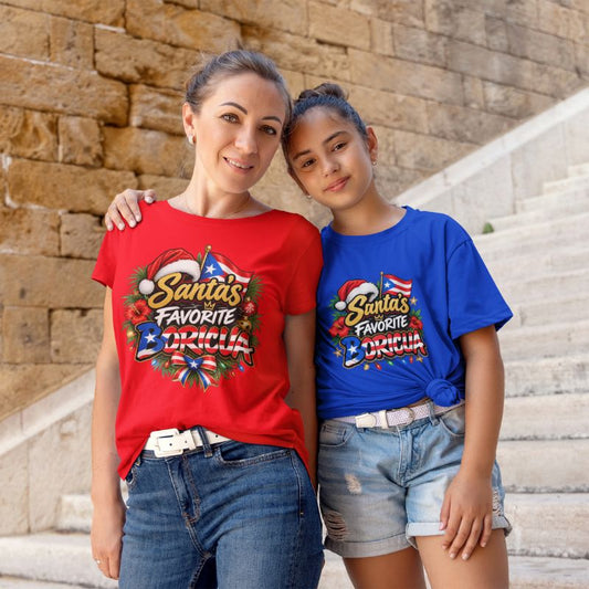 A woman and a young girl standing together on stone stairs, wearing matching graphic T-shirts. The woman wears a red shirt and the girl wears a blue shirt, both featuring a festive graphic that reads 'Santa's Favorite Boricua with a Santa hat, ornaments, and a Christmas tree motif.