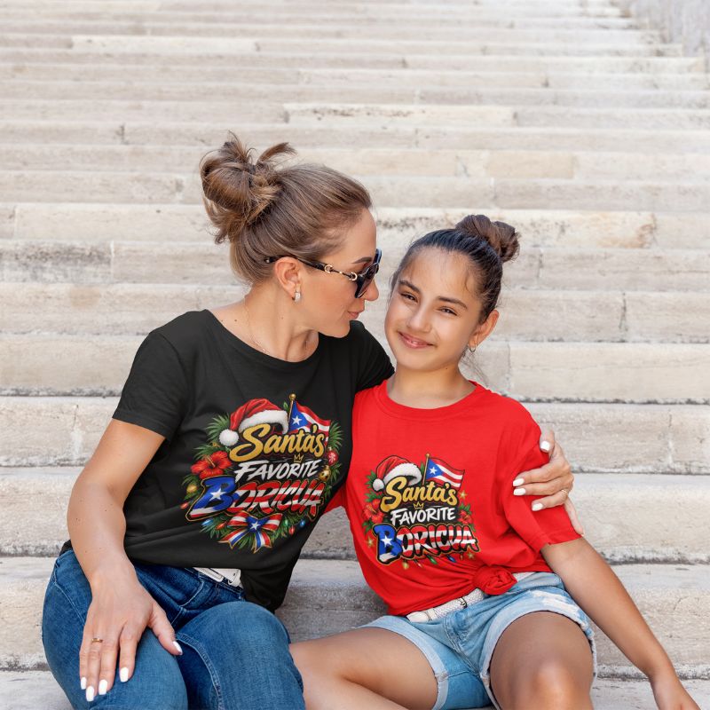 A woman and a young girl sitting on stone steps, both wearing matching graphic t-shirts with a colorful Santa-themed design that reads 'Santa's Favorite Boricua. The woman wears a black shirt, and the girl wears a red shirt.