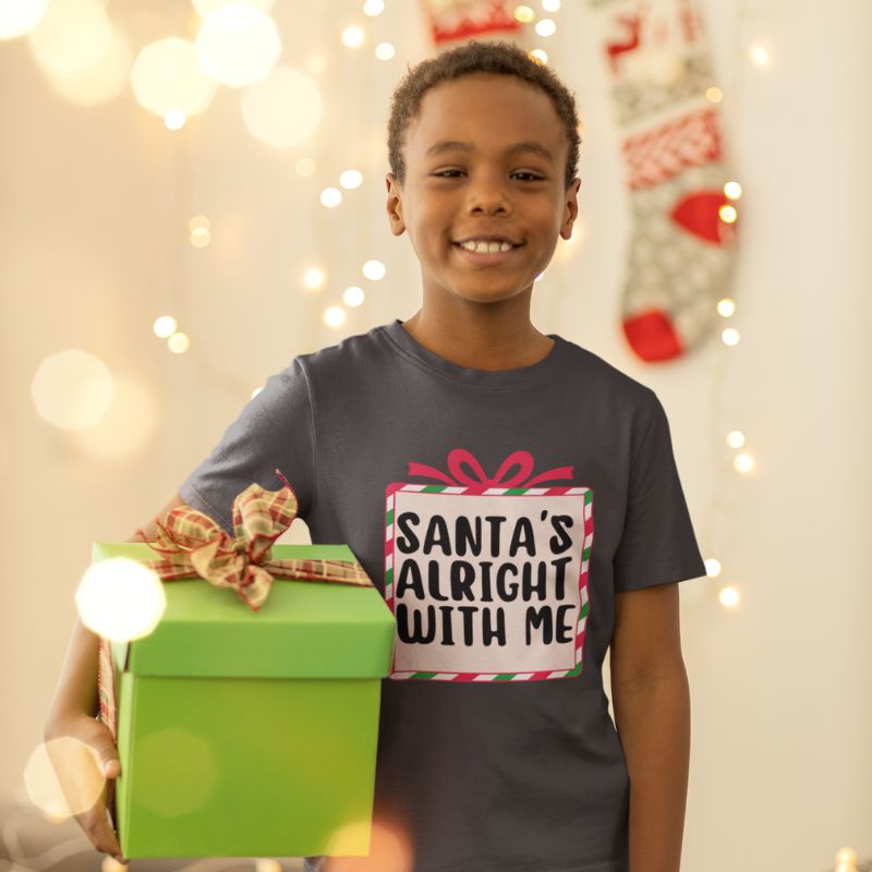 A smiling young boy wearing a dark gray graphic t-shirt that reads 'SANTA'S ALRIGHT WITH ME' in a gift box design, holding a bright green gift box with a plaid ribbon bow against a warm holiday background with string lights and a stocking.