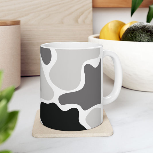 White mug with a black and gray abstract pattern on a coaster beside a fruit bowl.