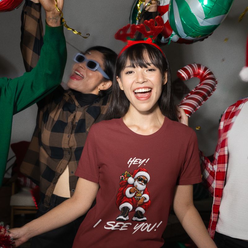 Smiling woman wearing a burgundy Hey! I See You! Santa graphic t-shirt at a festive gathering with holiday decorations.
