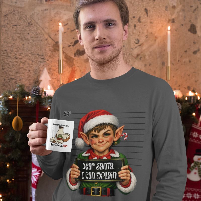 Man wearing a gray Dear Santa, I Can Explain long-sleeve shirt with an elf graphic, holding a white mug in a festive room with candles and holiday greenery.
