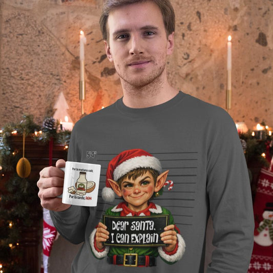 Man wearing a gray Dear Santa, I Can Explain long-sleeve shirt with an elf graphic, holding a white mug in a festive room with candles and holiday greenery.
