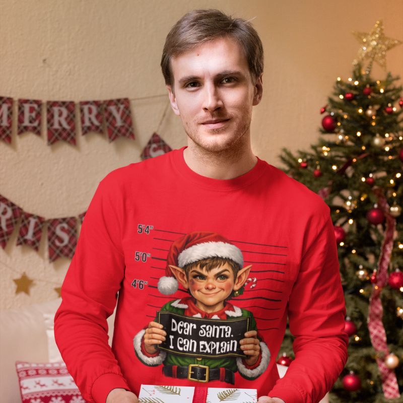 Man wearing a red Dear Santa, I Can Explain long-sleeve shirt featuring an elf holding a sign, standing beside a decorated Christmas tree.
