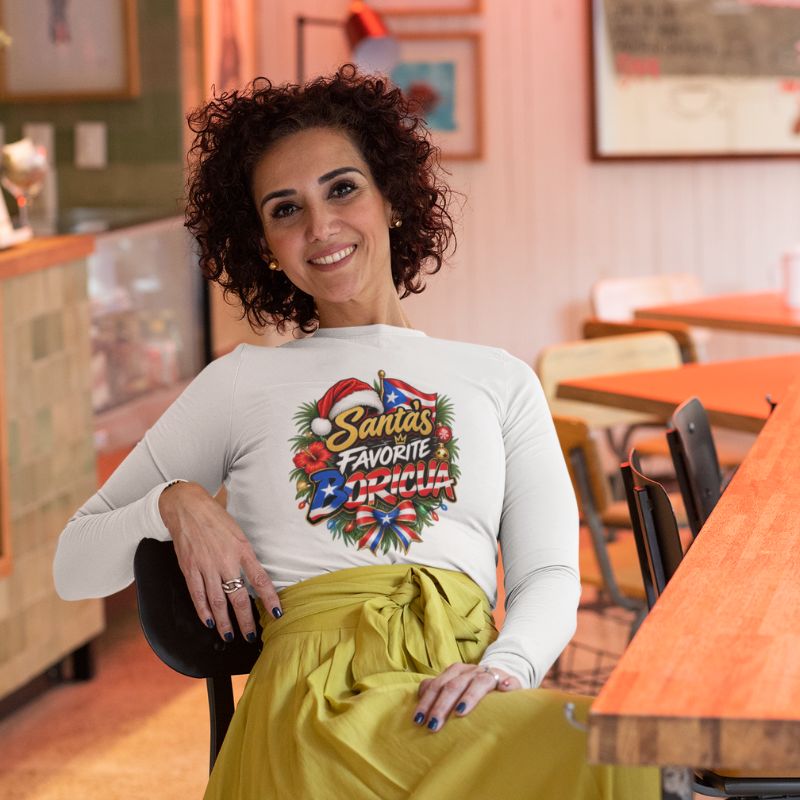 Woman wearing a white Santa’s Favorite Boricua Christmas long-sleeve shirt with yellow-green pants, seated in a café.
