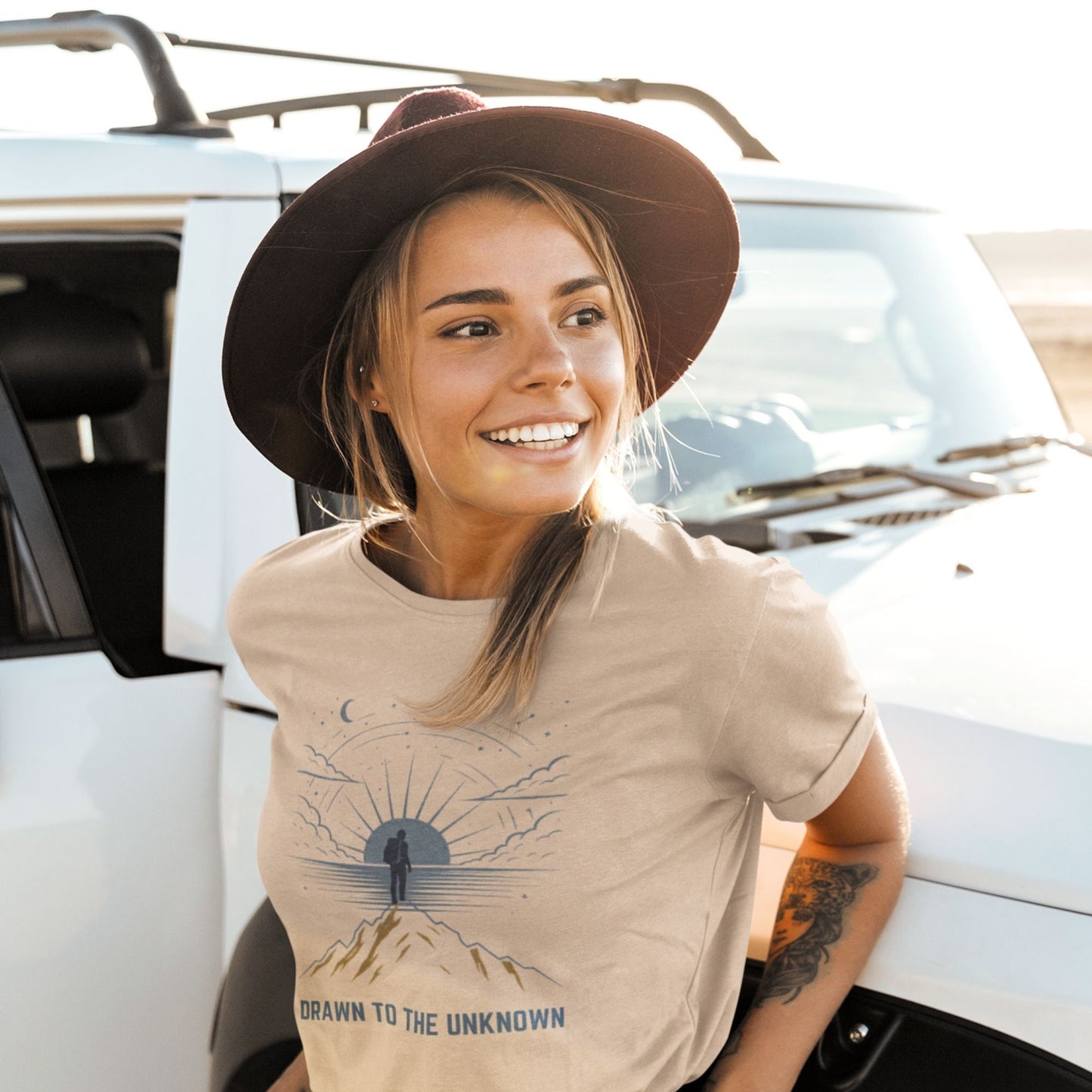  Woman in wide-brim hat wearing Drawn to the Unknown soft cream graphic tee leaning against white truck — boho adventure shirt

