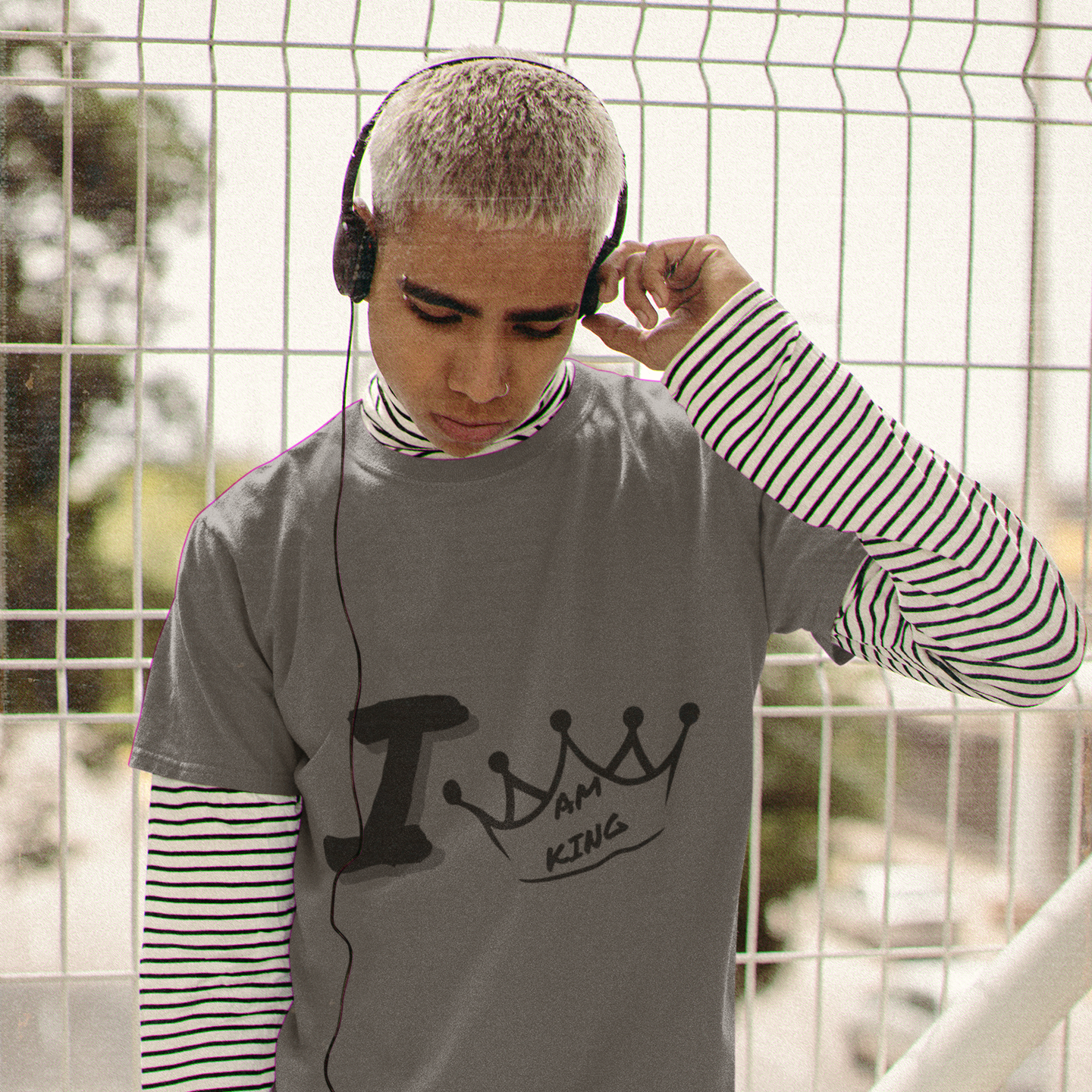 Person wearing a gray t-shirt with text, striped sleeves, and headphones, standing against a wire fence.