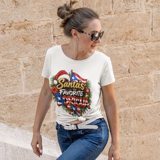 A woman wearing a white graphic t-shirt with a colorful 'Santa's Favorite Boricua design, paired with blue jeans, standing against a beige stone wall.