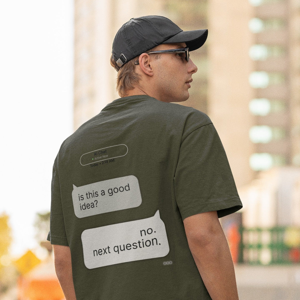 Man in cap and sunglasses showing back print of Is This a Good Idea AI chat graphic tee in military green
