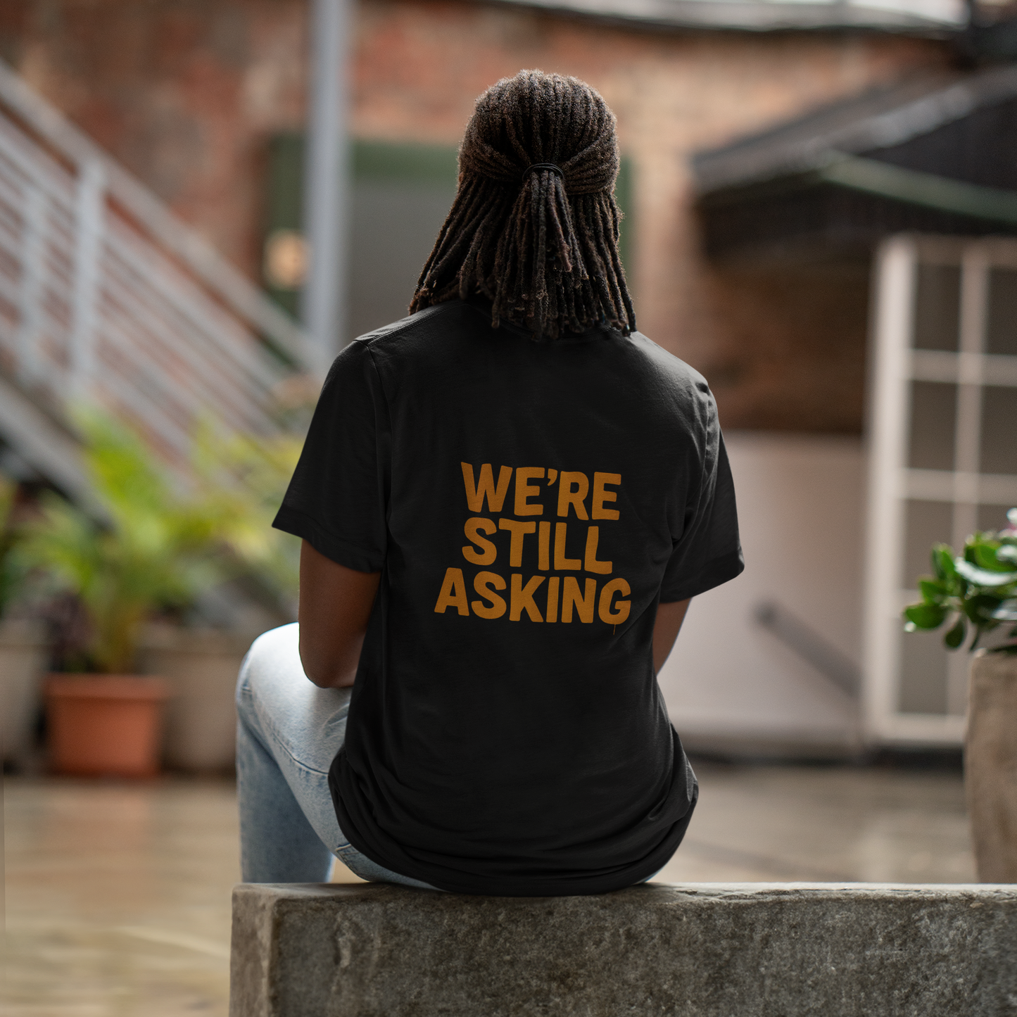 back view, outdoor wall): We're Still Asking back print graphic tee — gold lettering on black streetwear tee, rear view