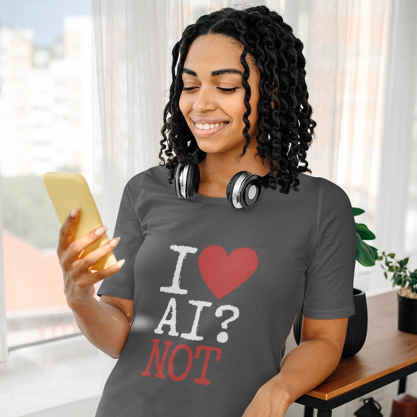 A woman with curly black hair stands indoors near a window, smiling at a phone while wearing a dark gray graphic T-shirt that reads 'I ♥ AI? NOT' in white and red text, with black-and-white headphones around her neck and blue jeans.