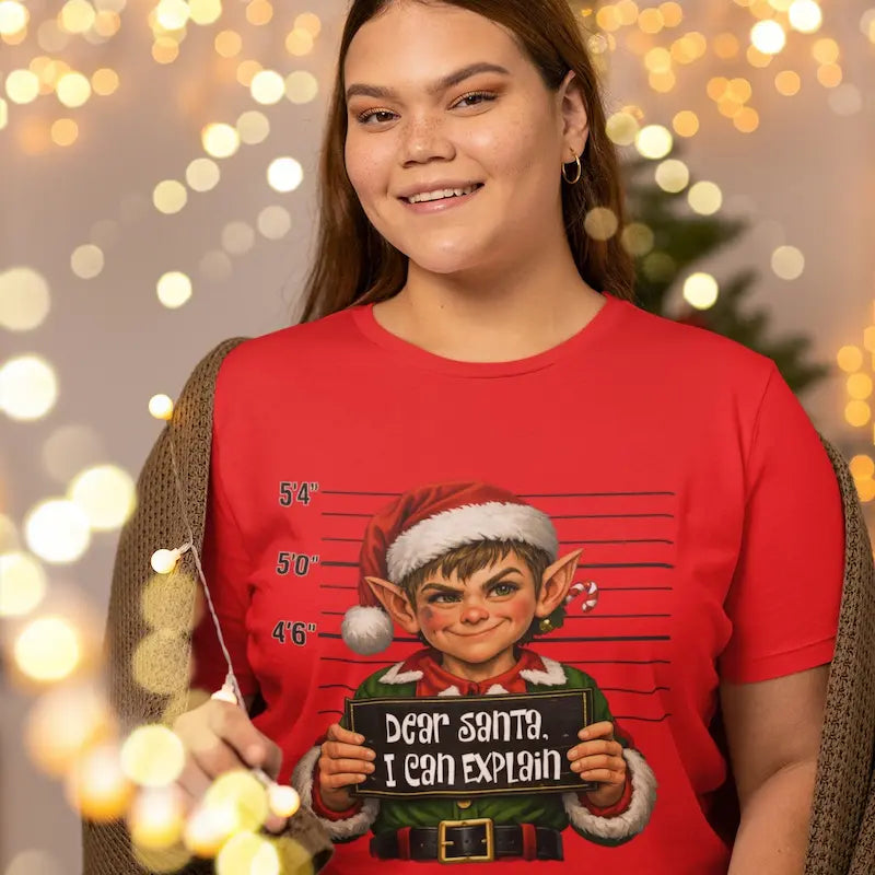 Woman wearing a red Dear Santa I Can Explain funny Christmas elf t-shirt in front of holiday lights