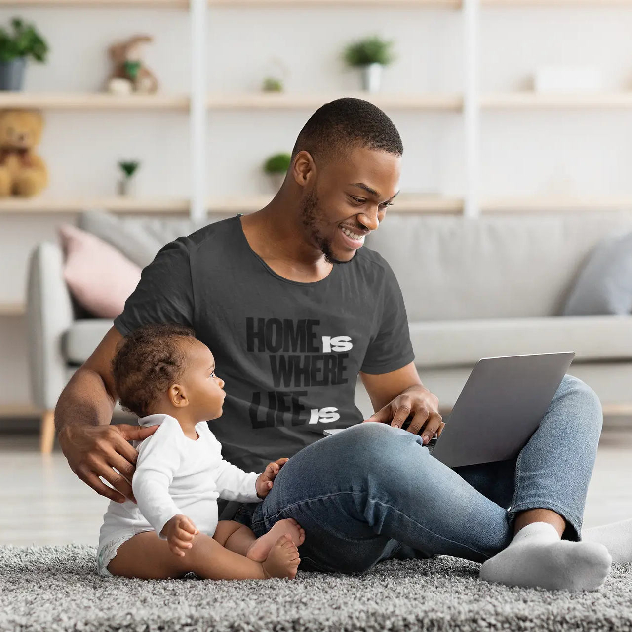 A smiling man sits on a gray rug in a bright living room, using a laptop while wearing a dark gray graphic T-shirt that reads 'HOME IS WHERE LIFE IS'. A baby in a white onesie sits beside him.