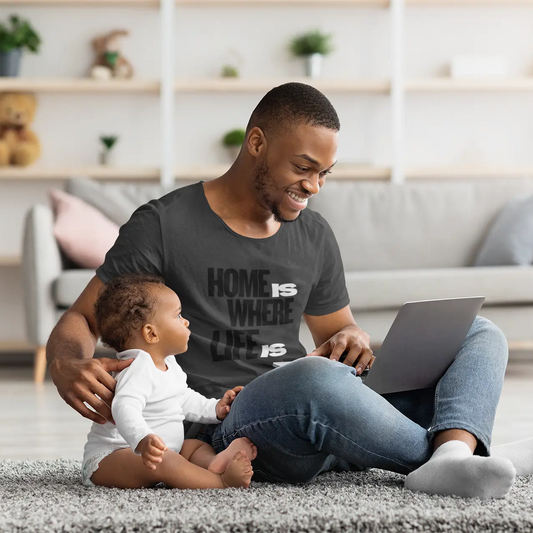 A smiling man sits on a gray rug in a bright living room, using a laptop while wearing a dark gray graphic T-shirt that reads 'HOME IS WHERE LIFE IS'. A baby in a white onesie sits beside him.