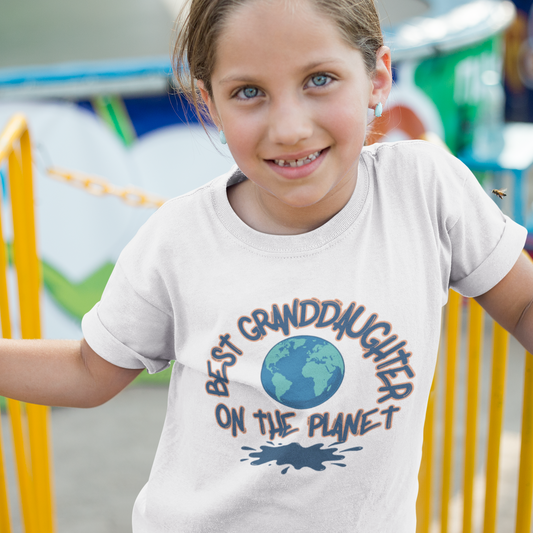 Young girl wearing a t-shirt with 'Best Granddaughter on the Planet' text at an outdoor playground.