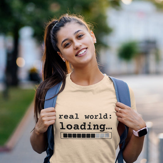 Soft cream Real World Loading graphic T-shirt worn by smiling female student with backpack outdoors — funny adulting streetwear tee Leela's Ink