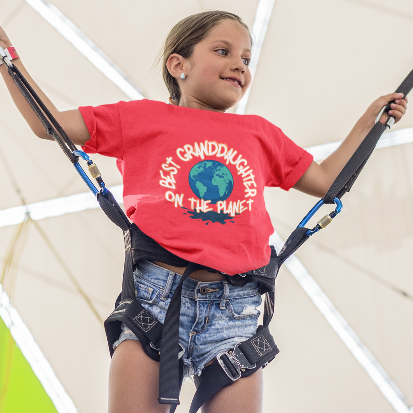 Child wearing a red shirt with text and denim shorts, standing on a trapeze with colorful geometric background