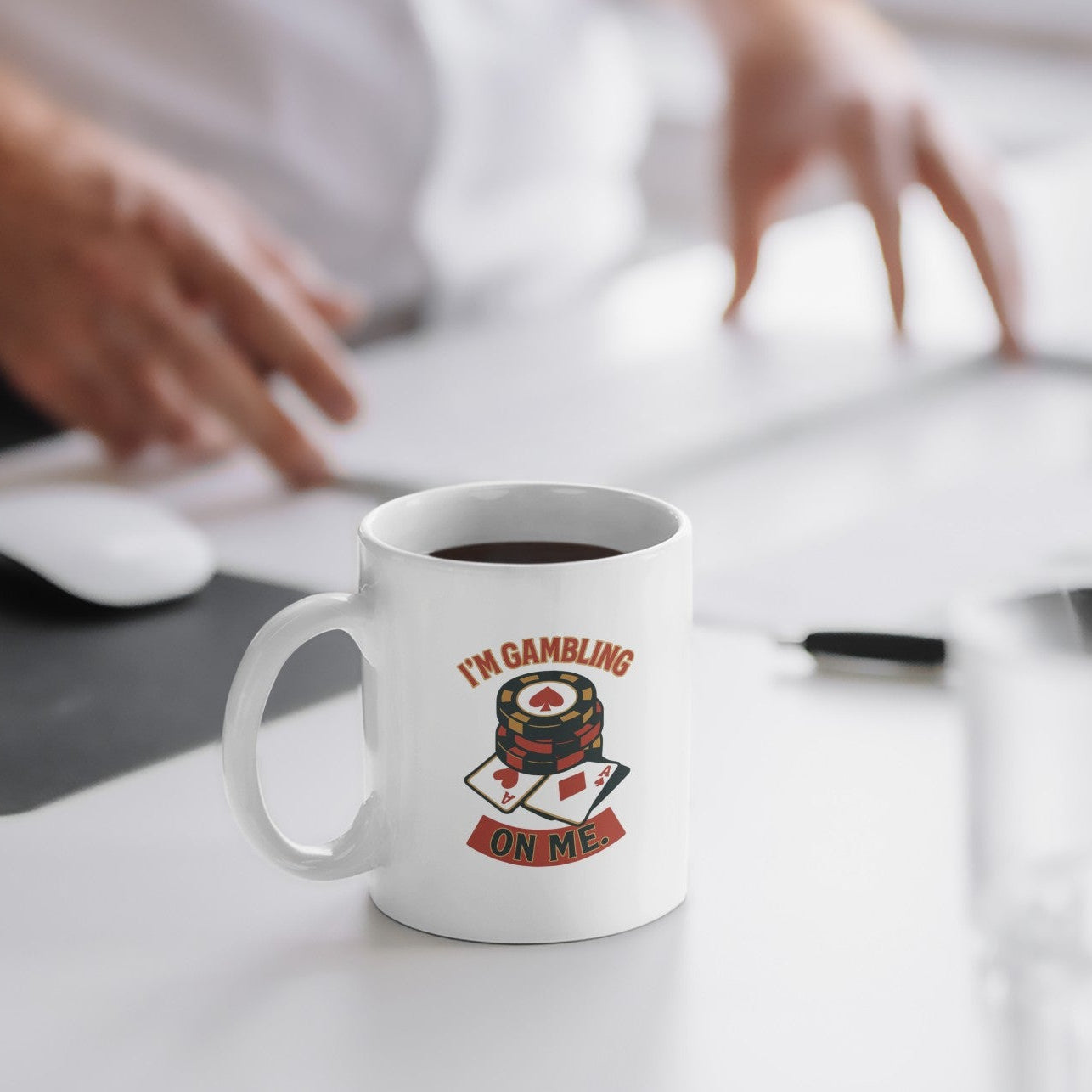 A white ceramic coffee mug with a 'I'M GAMBLING ON ME' graphic featuring a stack of poker chips and playing cards, sitting on a white desk in an office setting with a blurred person in the background.