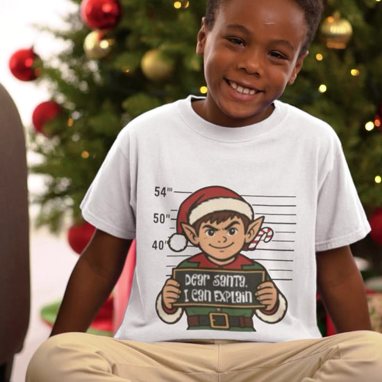 (video) Video of a smiling child sitting by a Christmas tree in a white Dear Santa I Can Explain kids elf t-shirt