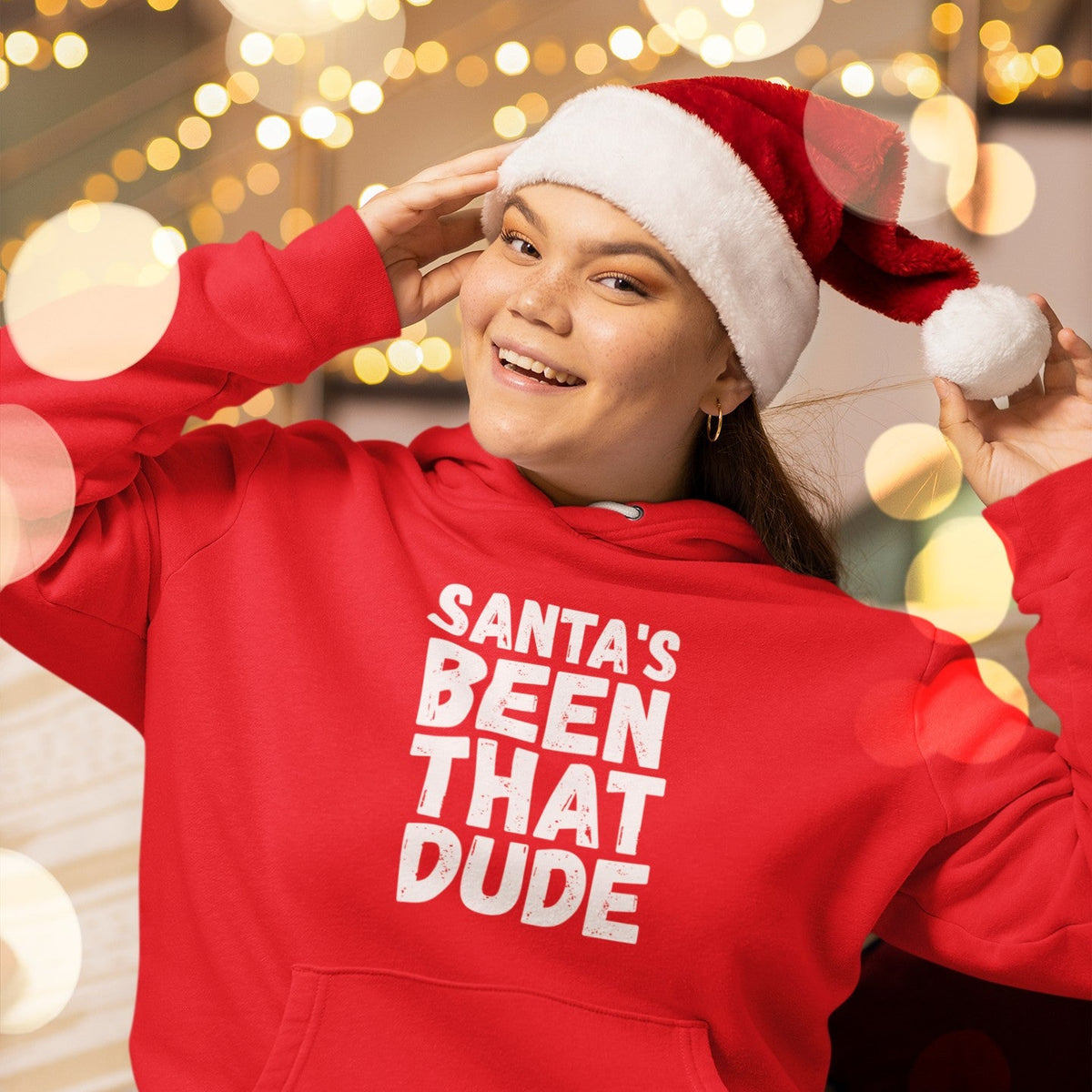 Person wearing a red “Santa’s Been That Dude” hoodie and Santa hat against holiday lights.