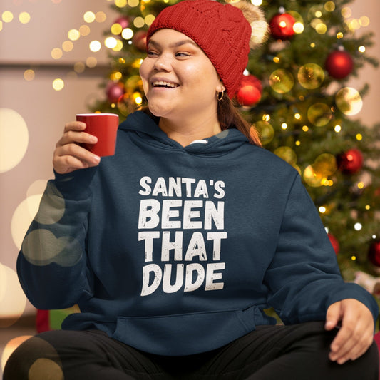 Person wearing a navy “Santa’s Been That Dude” hoodie and Santa hat beside a Christmas tree.