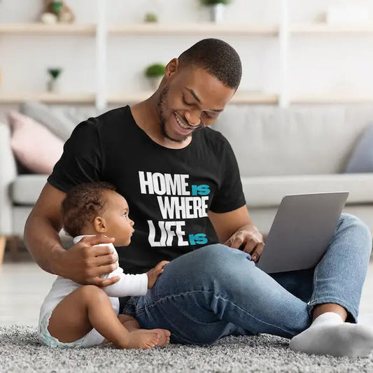A man sitting on a gray rug in a living room with a baby, wearing a black graphic t-shirt that reads 'HOME IS WHERE LIFE IS' in white and blue text. He is smiling and looking at a laptop.