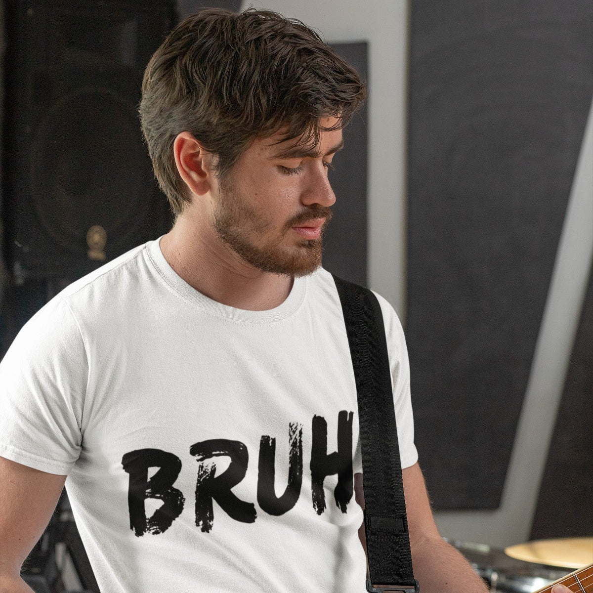 A man wearing a white graphic t-shirt with the word 'BRUH' printed in black distressed lettering, while playing an electric guitar in a studio setting.