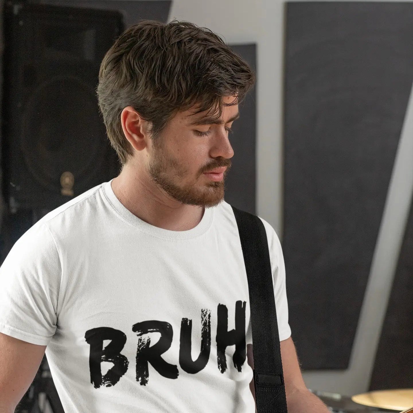 A man wearing a white graphic t-shirt with the word 'BRUH' printed in black distressed lettering, while playing an electric guitar in a studio setting.
