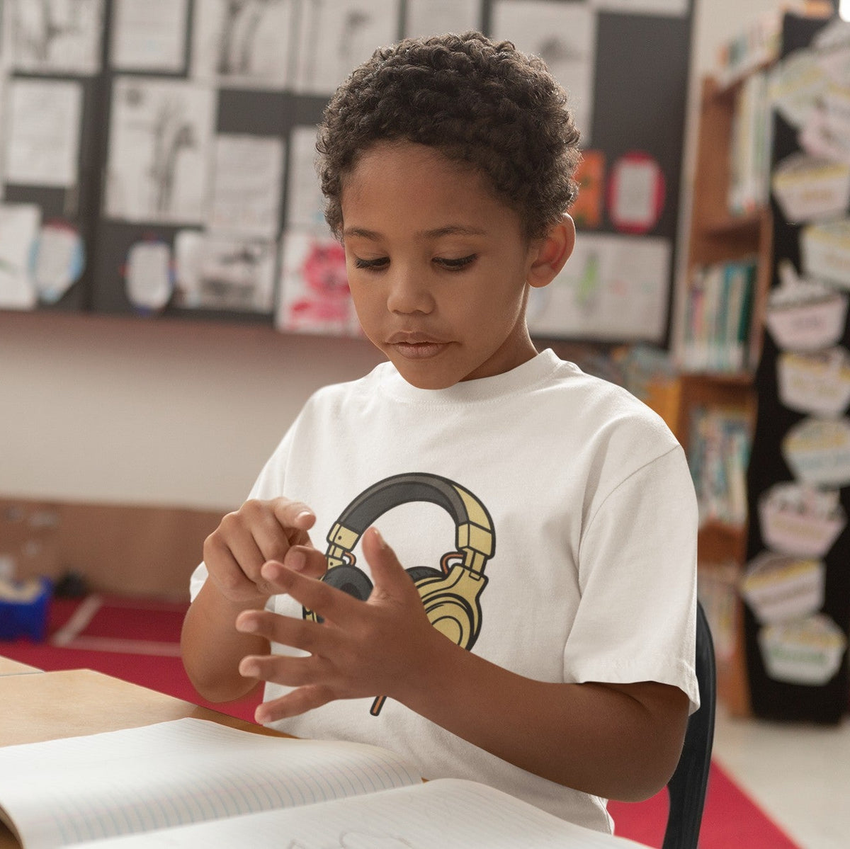 Young boy playing with a toy in a classroom wearing a white kids gold headphones graphic t-shirt — Leela's Ink

