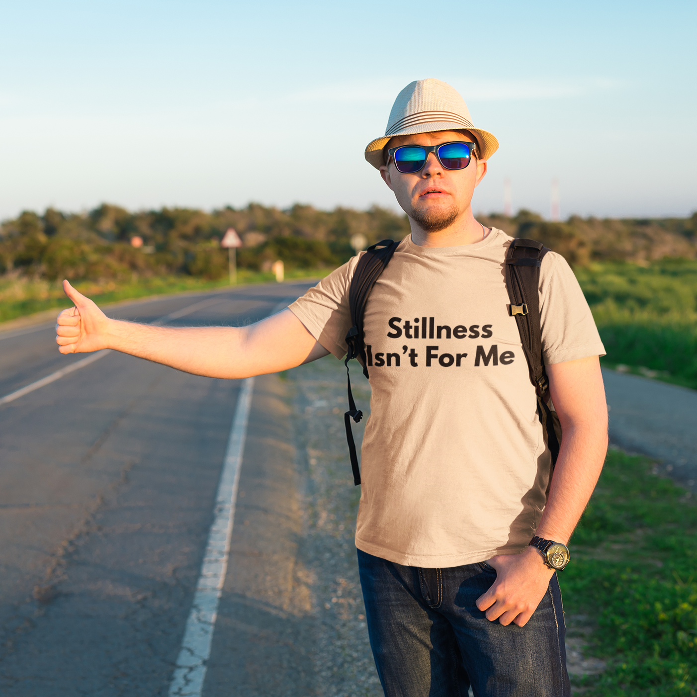 Man hitchhiking on open road wearing cream Stillness Isn't For Me graphic tee — adventure travel shirt

