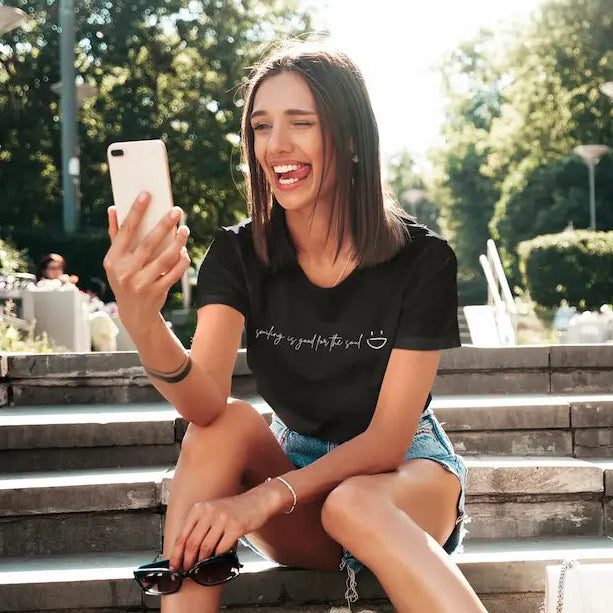 A woman sitting on outdoor stone steps in a sunny park, holding a white smartphone and wearing a black graphic T-shirt with the phrase 'writing is good for the soul' and a small smiley face.