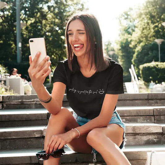 A woman sitting on outdoor stone steps in a sunny park, holding a white smartphone and wearing a black graphic T-shirt with the phrase 'writing is good for the soul' and a small smiley face.