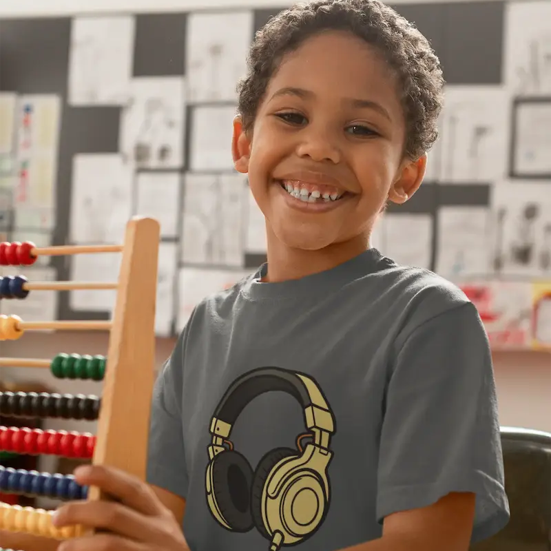 A smiling young boy with curly hair sits at a wooden table in a classroom, wearing a gray t-shirt with a graphic of headphones. He is holding a wooden abacus, with a chalkboard and educational posters in the background.
