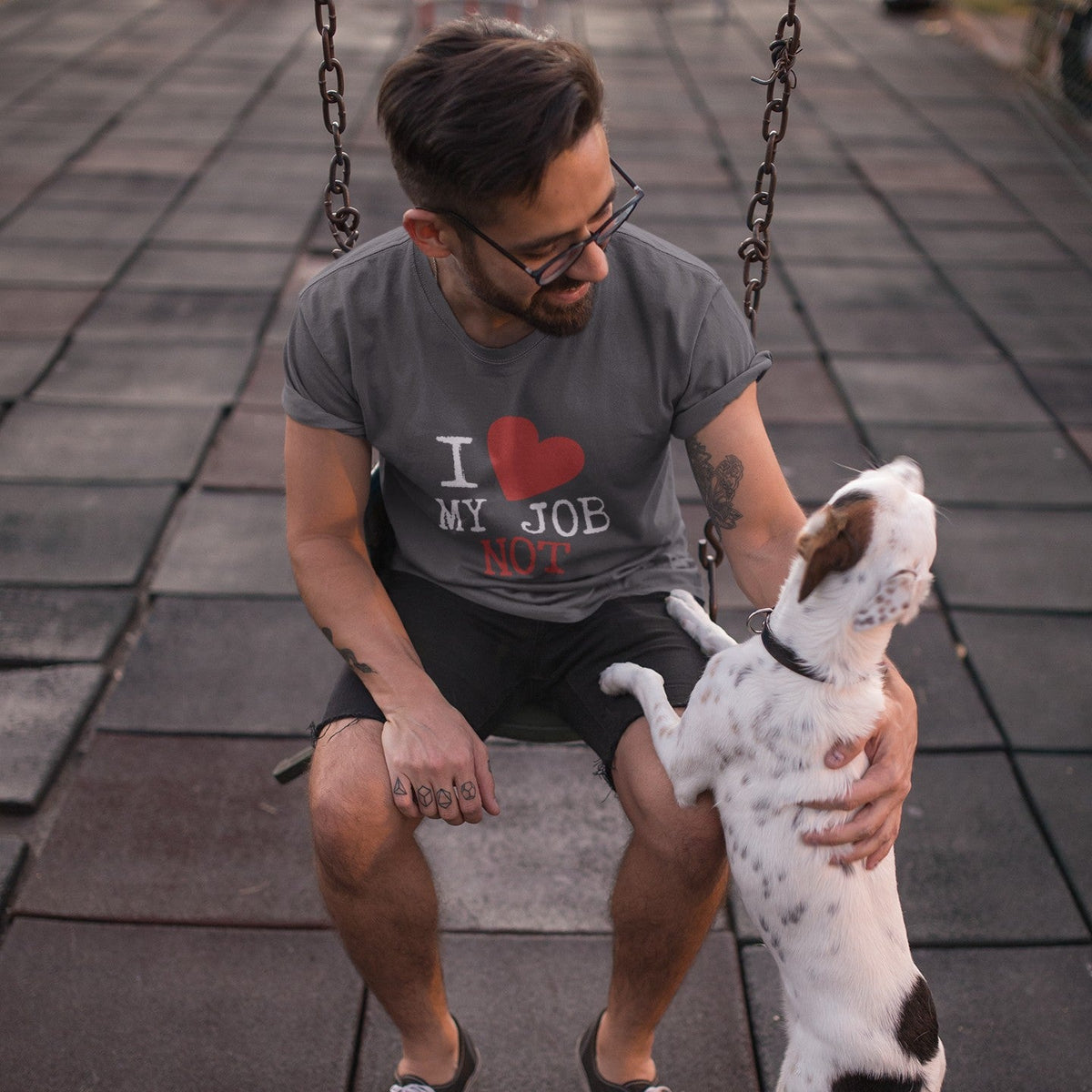 A man wearing a gray graphic t-shirt with 'I [heart] MY JOB NOT' printed on the front, sitting on a swing and petting a white dog with brown spots on a paved outdoor walkway.