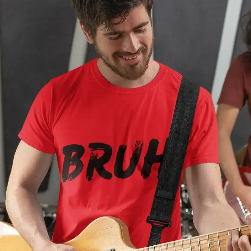 A man in a red t-shirt with 'BRUH' printed in black distressed lettering, playing a light-colored bass guitar in a music studio setting with another musician blurred in the background.