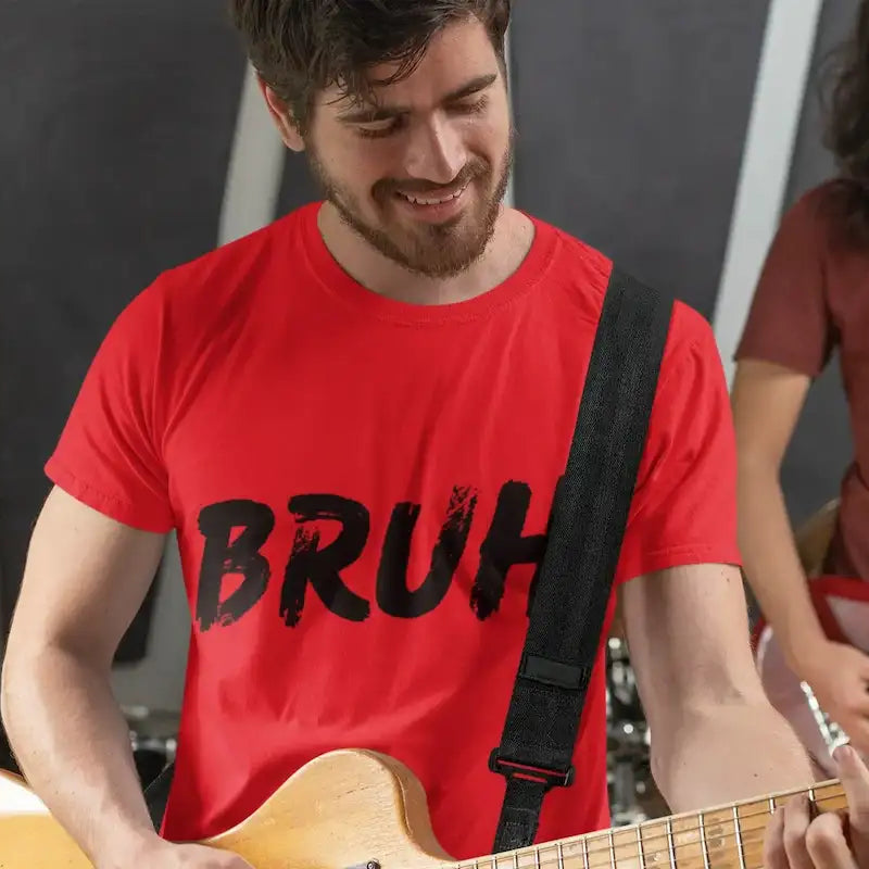 A man in a red t-shirt with 'BRUH' printed in black distressed lettering, playing a light-colored bass guitar in a music studio setting with another musician blurred in the background.