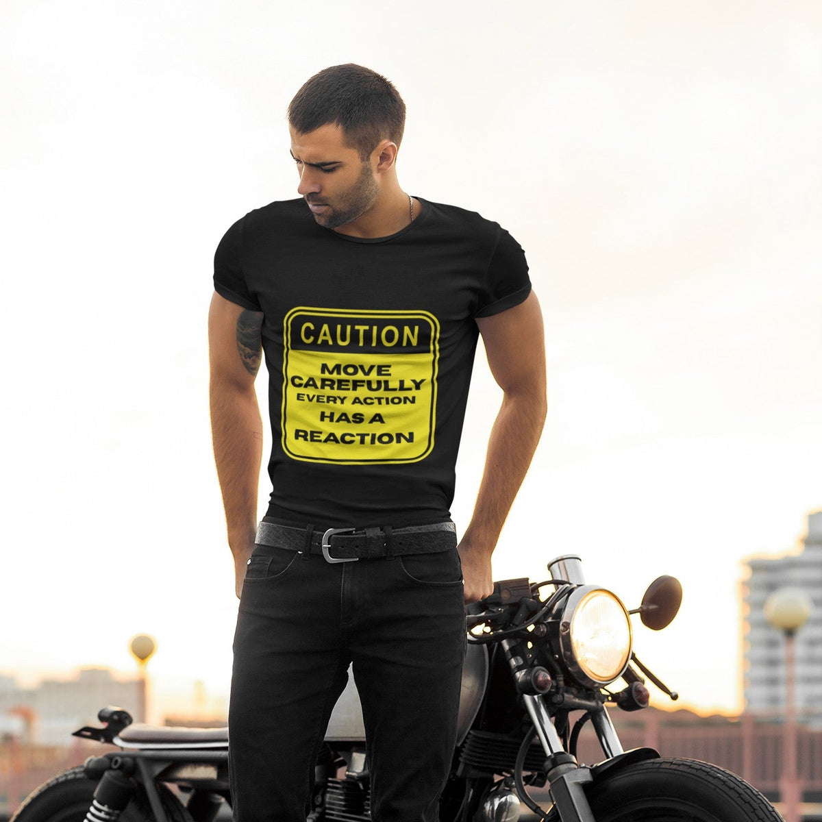 Man standing next to a motorcycle wearing a black Caution Move Carefully Every Action Has a Reaction graphic t-shirt